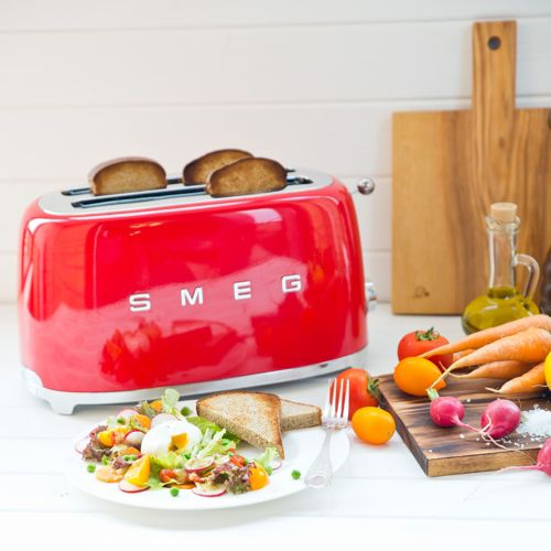 Ein roter Smeg-Toaster mit Toast, daneben ein Salat und Gemüse auf einem Holztisch.
