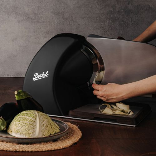 Una mano aziona un'affettatrice Berkel nera, tagliando zucchine. Cavolo e melanzane sono vicini.