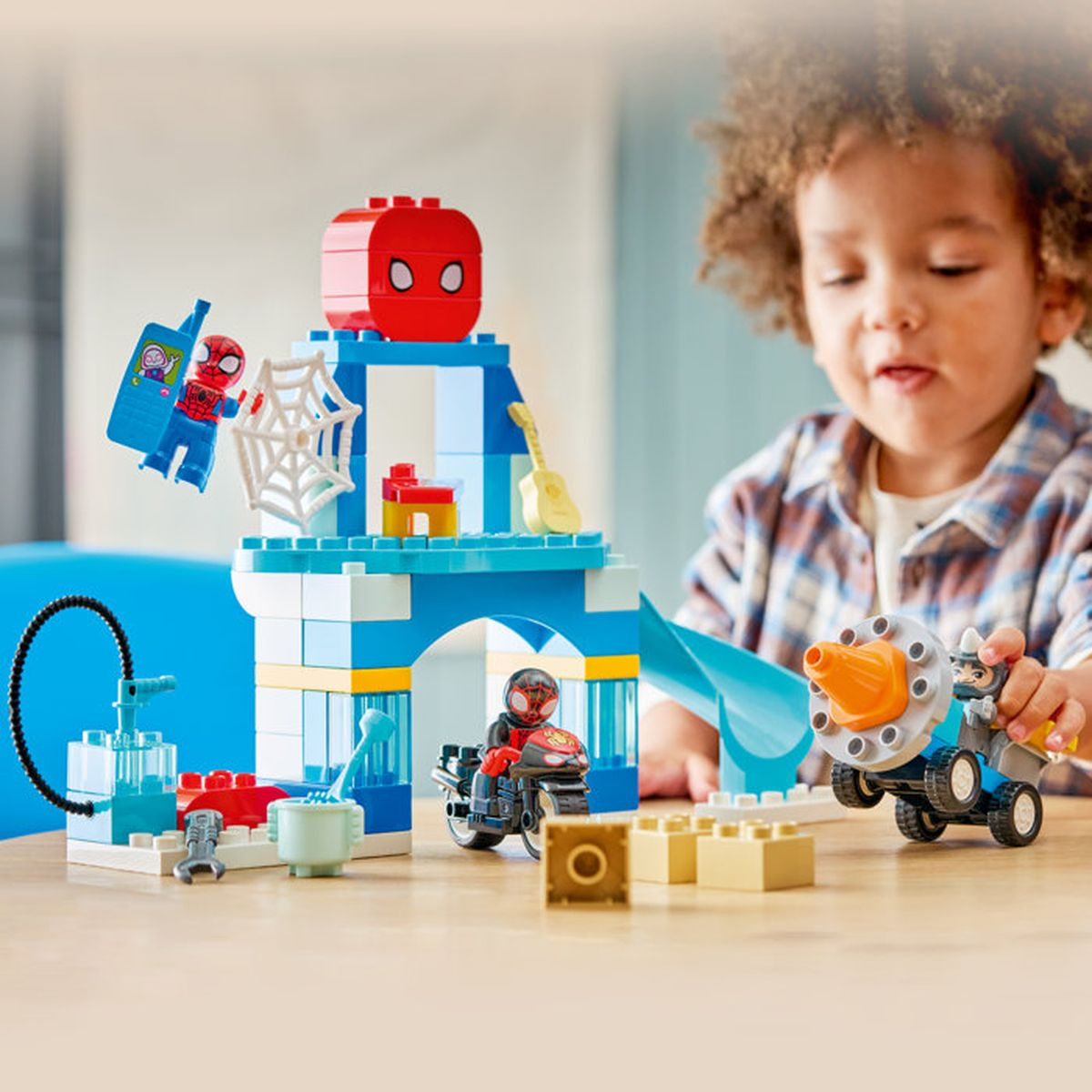 Bambino gioca con LEGO Spiderman: edificio blu, figure e veicoli su un tavolo.