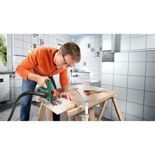 Mann mit orangefarbenem Pullover schneidet Holz mit Kreissäge in gefliestem Raum.