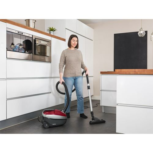 Una mujer aspira el suelo de una cocina moderna con una aspiradora roja y gris.