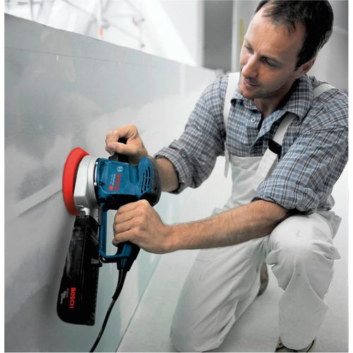 Hombre lija pared con lijadora Bosch azul. Viste overol y camisa a cuadros.