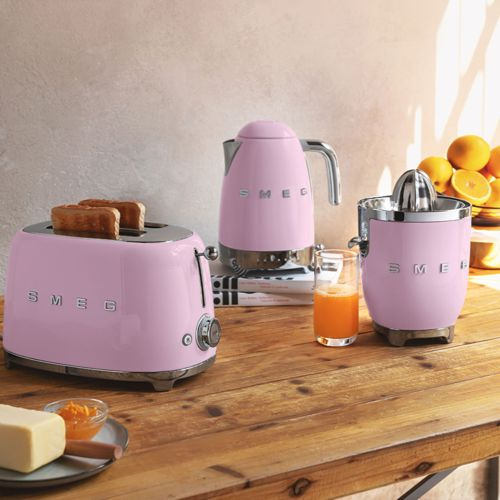 Üç pembe SMEG mutfak aleti: ekmekli tost makinesi, su ısıtıcısı ve narenciye sıkacağı, ahşap masa üzerinde.