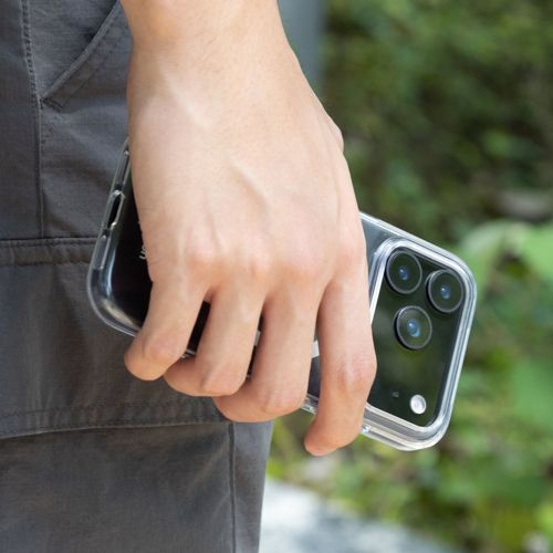 Una mano tiene una custodia trasparente per telefono con tre fotocamere. Pantaloni grigi visibili.