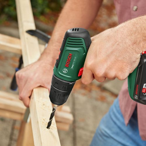 Mężczyzna wierci w kawałku drewna zieloną wiertarką akumulatorową Bosch.