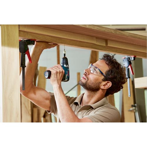 Mann mit Schutzbrille bohrt Holzbalken. Zwingen halten Holz fest.