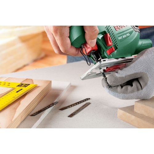 Persona usa sierra de calar, hojas de sierra, madera y escuadra en mesa.