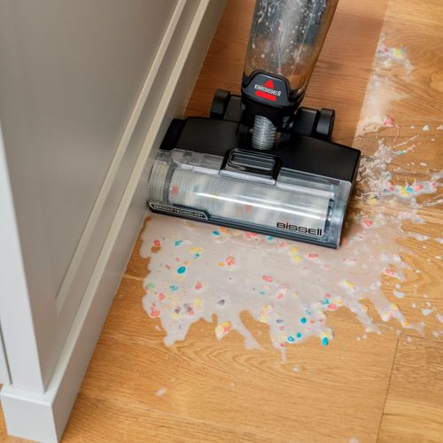 Un aspirapolvere bagnato/asciutto pulisce briciole colorate e liquidi su un pavimento in legno.