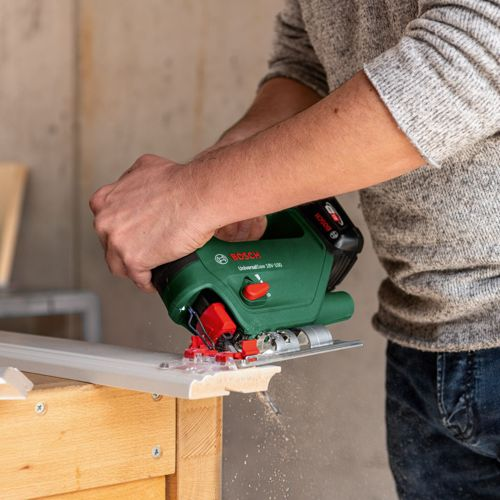 Eine Person schneidet mit einer grünen Bosch Stichsäge weiße Zierleisten auf Holz.