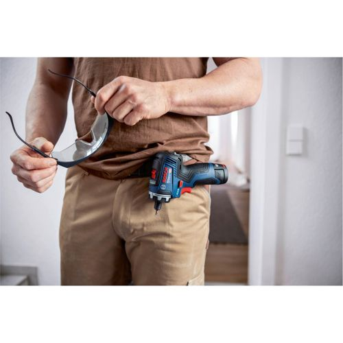 Homme en chemise marron et pantalon cargo tient lunettes de sécurité, perceuse à la ceinture.