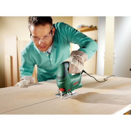 Man met veiligheidsbril gebruikt groene decoupeerzaag op houten plank.