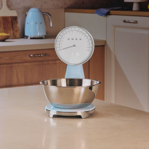 Una báscula de cocina azul con un bol plateado en una encimera. Una tetera azul al fondo.