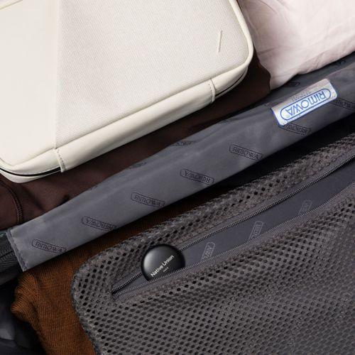 Eine weiße Tasche liegt auf einem gepackten Rimowa-Koffer mit einem kleinen schwarzen Tracker darin.