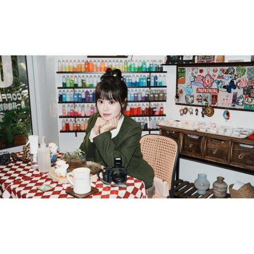 Une femme est assise à une table recouverte d'une nappe à carreaux devant une étagère de bouteilles colorées.