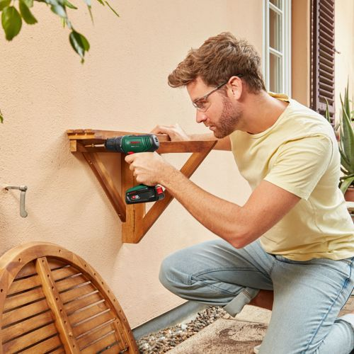 Uomo avvita mensola di legno al muro con trapano. All'aperto.