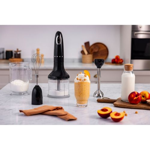 Een zwarte SMEG staafmixer met accessoires, een perzik smoothie en fruit op een marmeren aanrecht.