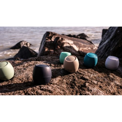 Six enceintes Bluetooth colorées posées sur des rochers près de l'eau.
