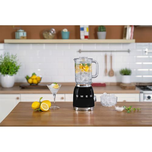 Buz, limon ve portakal dilimleri olan siyah bir Smeg blender ahşap masada duruyor.