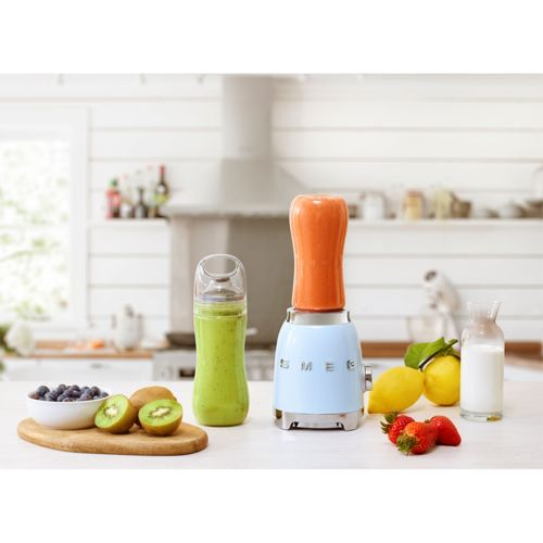 Ein hellblaues SMEG-Mixgerät mit einer orangefarbenen Tasse daneben. Links ein grüner Smoothie und rechts Früchte.