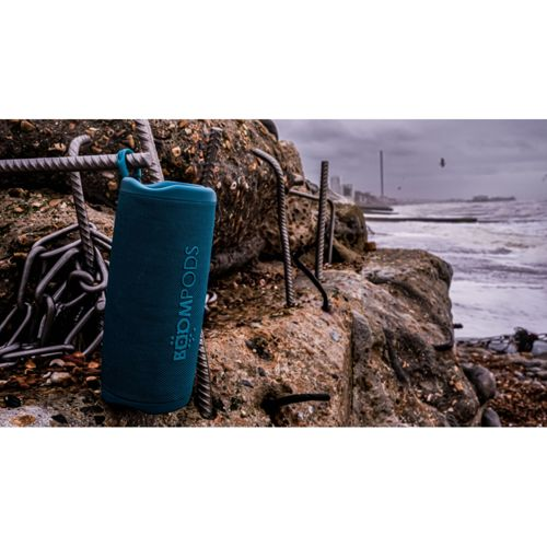 Un altoparlante blu Boompods è appeso a una fune metallica vicino a rocce sulla spiaggia.