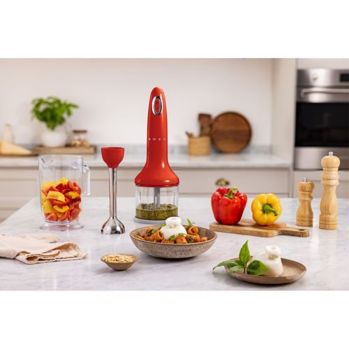 Un frullatore a immersione rosso vicino a una caraffa di frutta, pasta e macinapepe.