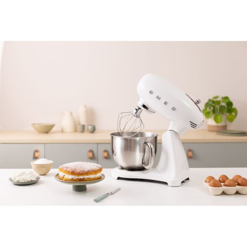 Eine weiße Küchenmaschine mit Schneebesenaufsatz steht neben einem Kuchen auf der Theke.