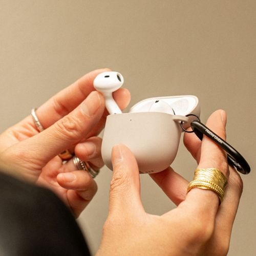 Una mano inserisce un auricolare wireless bianco in una custodia di ricarica beige. Anelli dorati adornano le dita.