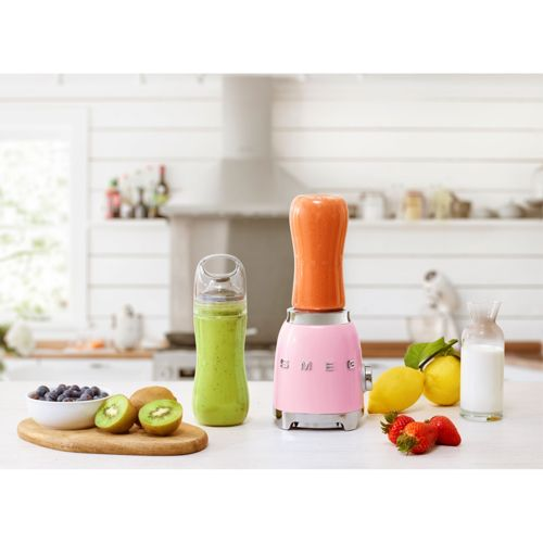 Un frullatore rosa con bicchiere per frullato arancione, accanto a frullato verde, bacche, kiwi, limoni, fragole e latte.
