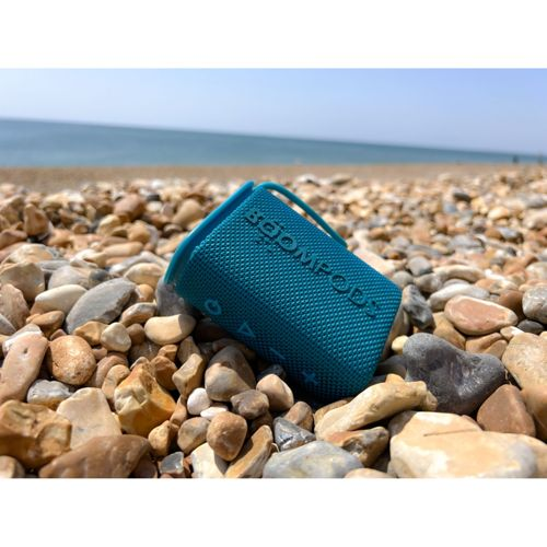 Une enceinte bleue repose sur une plage de galets avec la mer en arrière-plan.