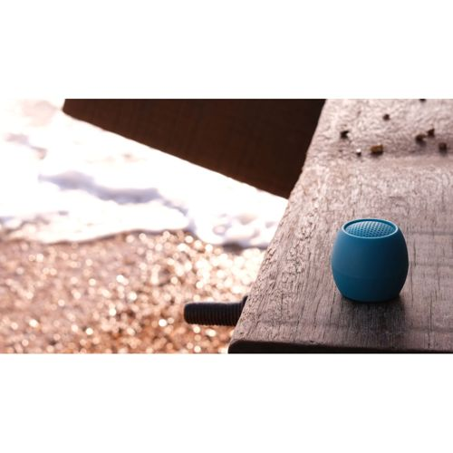 Un haut-parleur bleu sur du bois avec plage et mer à l'arrière-plan.