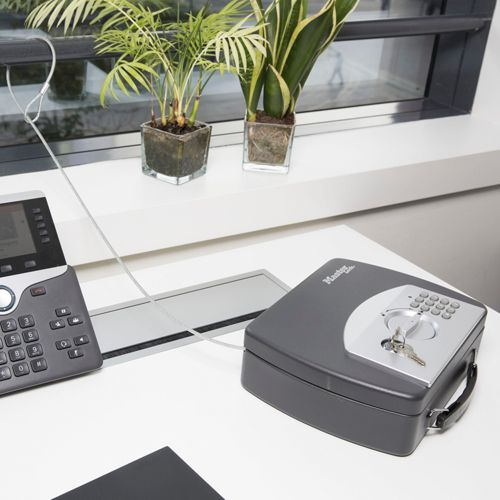 Un teléfono de oficina gris y una caja fuerte cerrada sobre un escritorio blanco, con plantas al fondo.