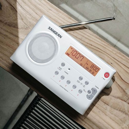 A white Sangean radio sits on a wooden table, displaying 100.7 FM and 8:00 AM.