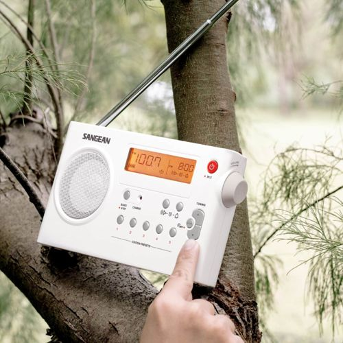 A white Sangean radio with antenna leans on a tree trunk. A hand presses a button.