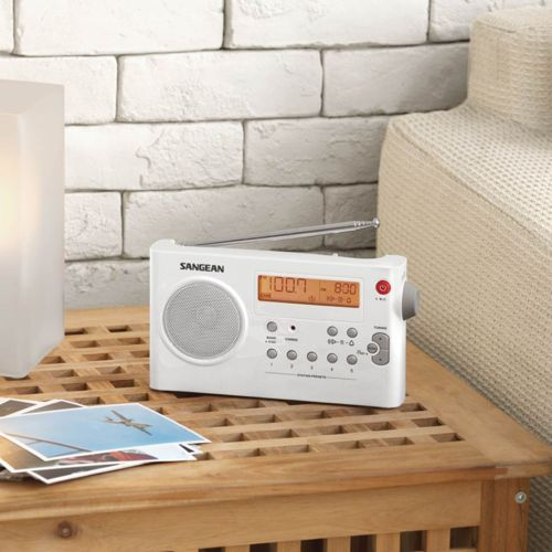 A white Sangean radio sits on a wooden table next to photos and a lamp.