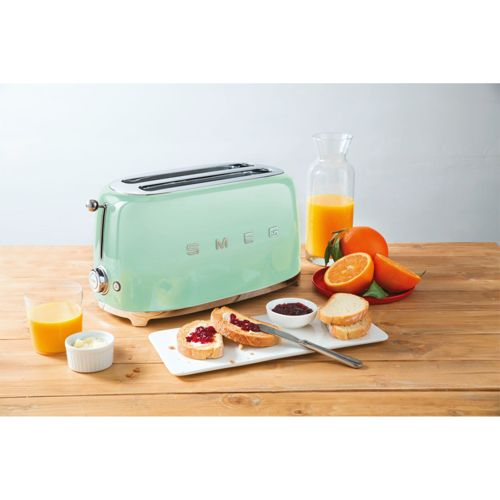 Una tostadora Smeg verde pastel con tostadas, mermelada, mantequilla, zumo de naranja y naranjas.