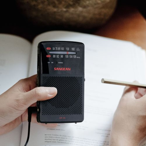 Black Sangean radio with AM/FM dial, held in a hand over a book.