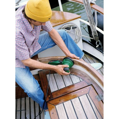 Mann schleift Holzboot mit Exzenterschleifer, trägt gelbe Mütze und Jeans.