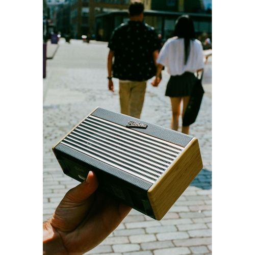 Une main tient une petite enceinte noire aux côtés en bois. Un couple marche en arrière-plan.