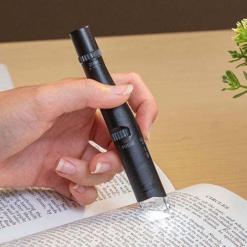 Una mano tiene una penna d'ingrandimento nera con ghiere di zoom e messa a fuoco su un libro aperto.