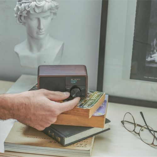 Een hand stelt een houten radio op boeken bij. Een buste en bril op de achtergrond.