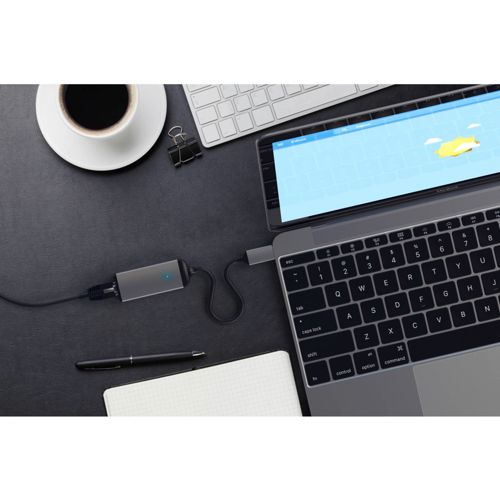 Un laptop è collegato a un hub grigio. Caffè, taccuino e penna sono nelle vicinanze.