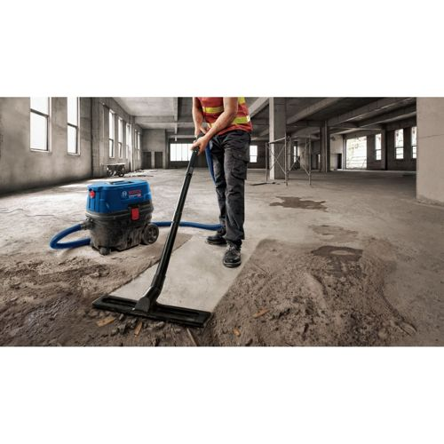 Operaio aspira detriti con aspirapolvere industriale Bosch blu in edificio vuoto.