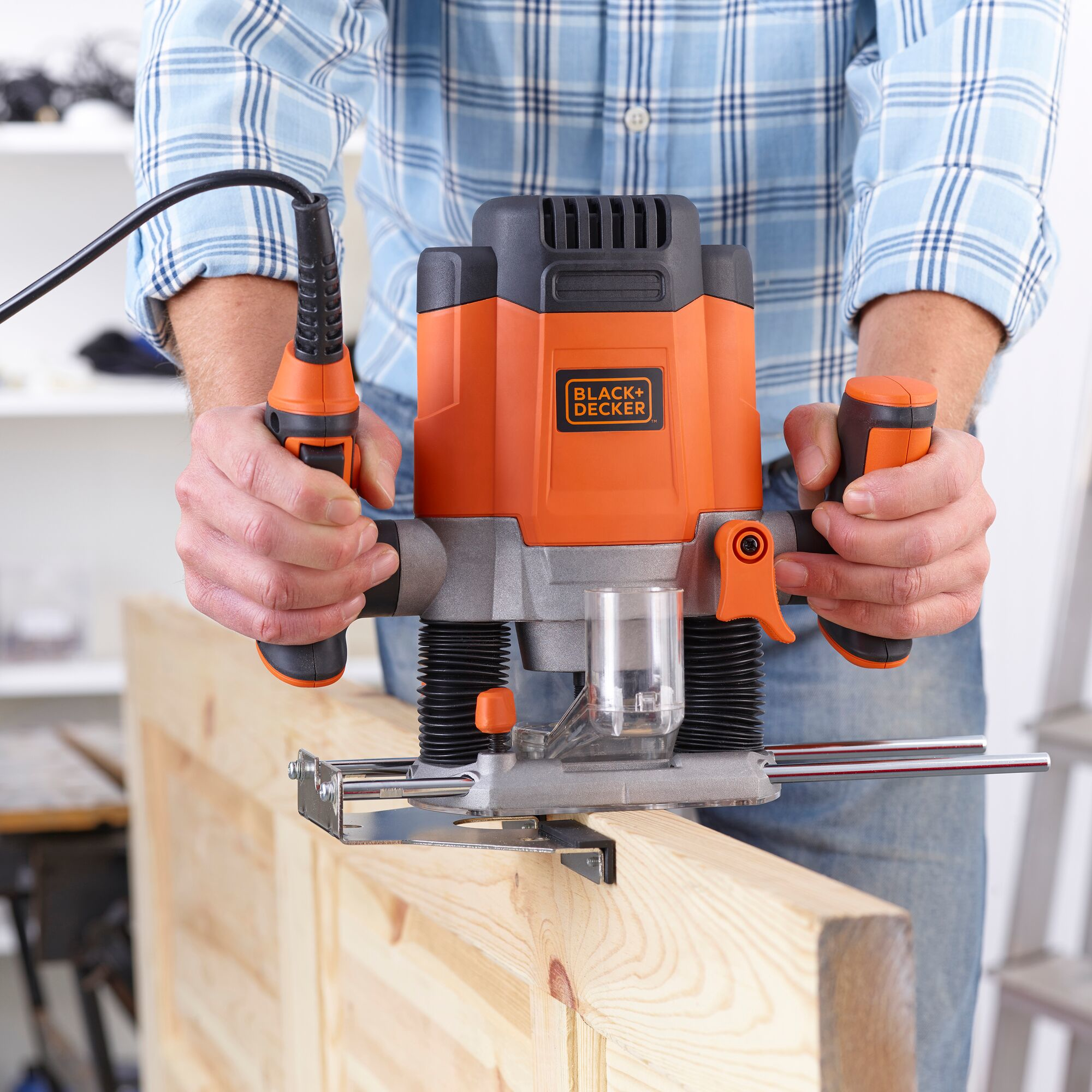 Hombre usa una fresadora en madera. Herramienta negra-naranja, camisa de cuadros azules.