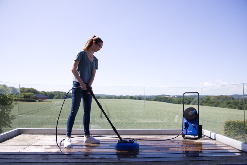 Donna pulisce terrazzo in legno con idropulitrice, macchina blu, campo dietro.