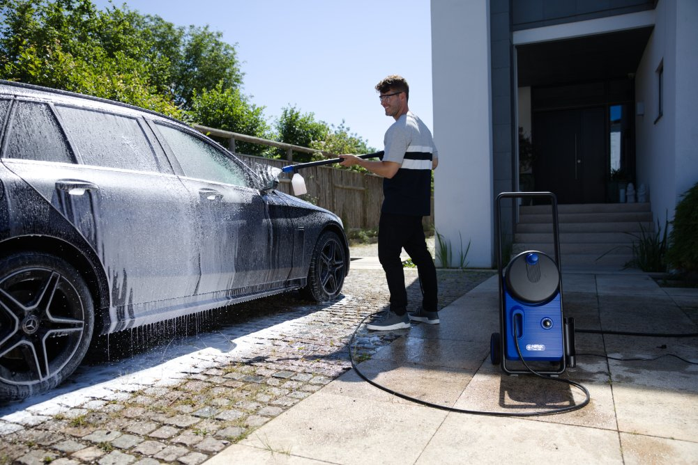 Un uomo lava un'auto con un'idropulitrice davanti a una casa moderna.