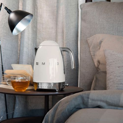 Een witte Smeg waterkoker staat naast een boek en een kop thee op een bijzettafel.