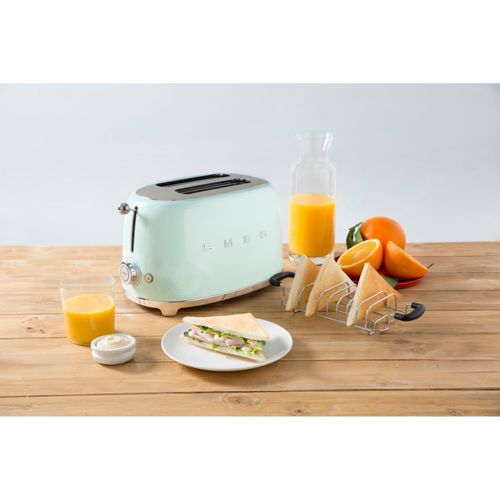 Ein mintgrüner SMEG-Toaster steht auf einem Holztisch mit Toast, Saft und Orangen.