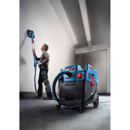 Mann schleift Wand mit langem Gerät, blauer Industriestaubsauger daneben.