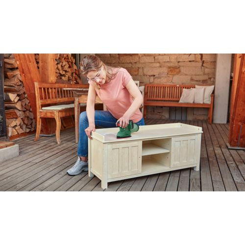 Frau schleift Holzschrank mit grünem Schleifgerät auf einer Holzterrasse.