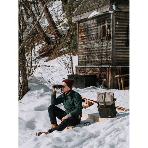 Mann mit Fernglas sitzt im Schnee vor Holzhütte.
